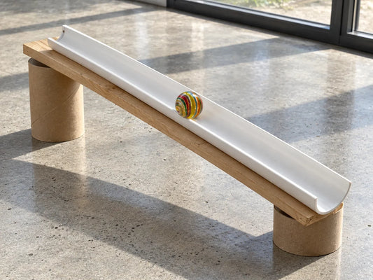 Bille de verre à rayures multicolores dans une gouttière blanche posée en pente sur une planche de bois et deux rouleaux de carton, sur un sol de béton ciré ensoleillé