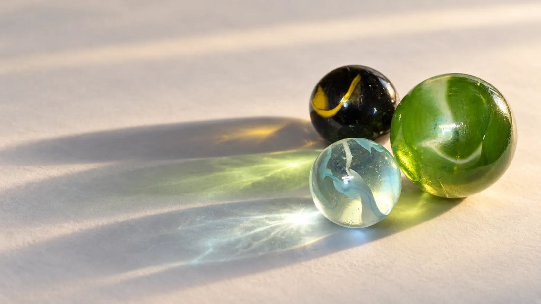 Trois billes de verre MesBilles posées sur une surface claire à contre-jour, projetant chacune une traînée de lumière colorée